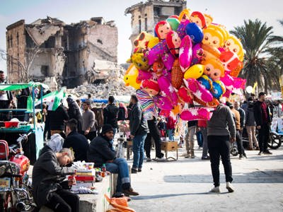 A destroyed building stands in the distance as crowds walk through the Alepp Citadel