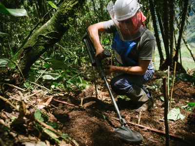 A deminer wearing PPE crouches between plants and uses their metal detector just above the soil