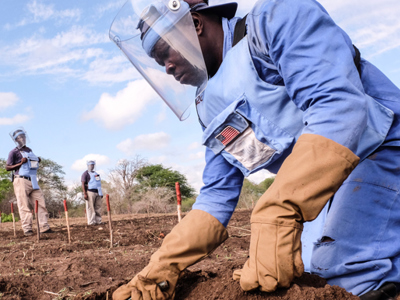 A man wearing PPE whilst kneeling on the ground and manually demining fields in Zimbabwe