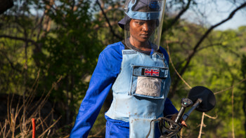 A deminer wearing protective gear walks through a field whilst holding a detector