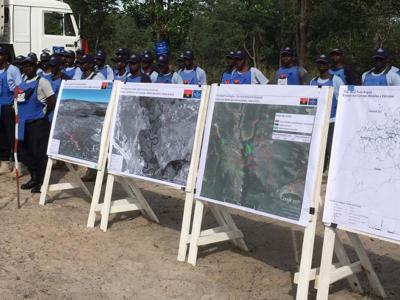 A large group of HALO Trust deminers stand behind boards displaying images and maps highlighting the contmination following the battle of the Cuito Cuanavale