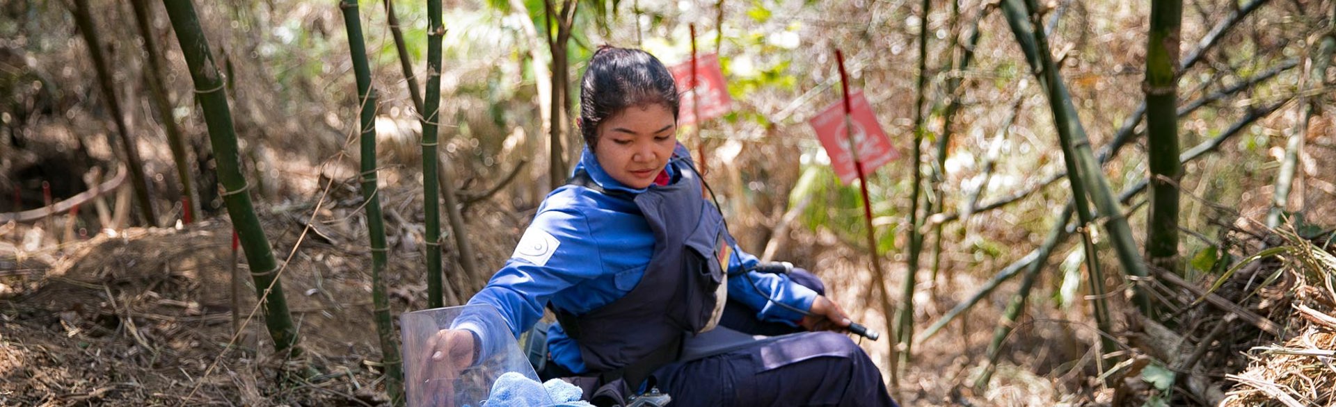 Female deminer sits on a forest hill with mine warning signs and her metal detector