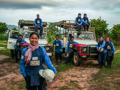 A group of deminers in Cambodia smile in front of their vehicles wearing PPE