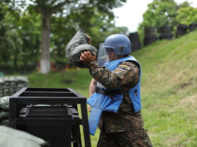 A HALO Trust Weapons and Ammunition technician holds a large sandbag whilst wearing a protective helmet and visor