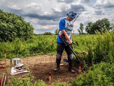 A deminer wearing PPE uses a metal detector to scan the shrubs amongst mine markers