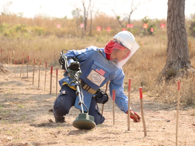 A deminer wearing protective gear kneels on mine marked ground while holding a detector