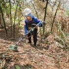 A female deminer wears PPE and holds a metal detector in a Cambodian forest in search of mines