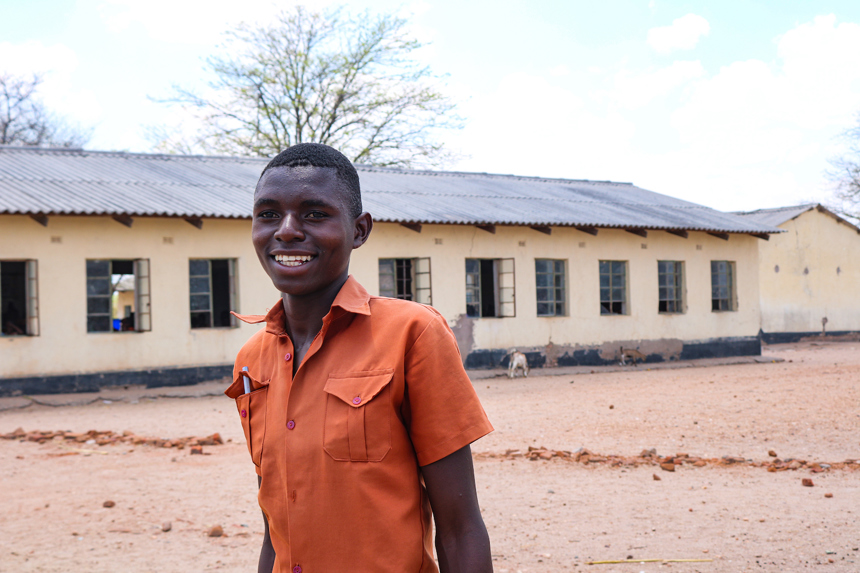 A young man wearing an orange shirt smiles in front of a building 