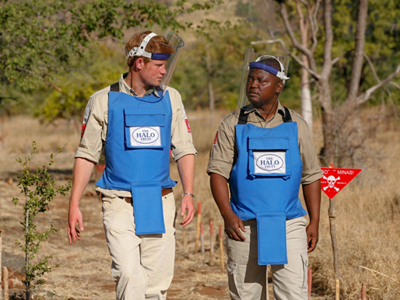 Prince Harry and Angolan HALO staff walk through a minefield wearing PPE